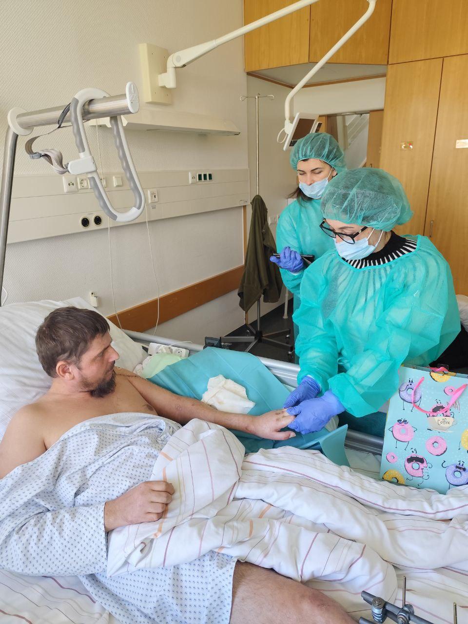 Zwei Helferinnen in grünen Schutzkitteln besuchen im Krankenhaus einen verwundeten Patienten im Bett und bringen ihm ein Geschenk.